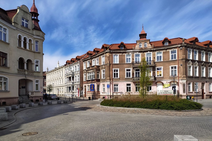 Cichy rynek w Gdańsku z brukowanymi uliczkami, zabytkowymi budynkami z dachami pokrytymi czerwoną dachówką oraz niewielkim rondem z zielenią pośrodku, a nad nim częściowo zachmurzone błękitne niebo.