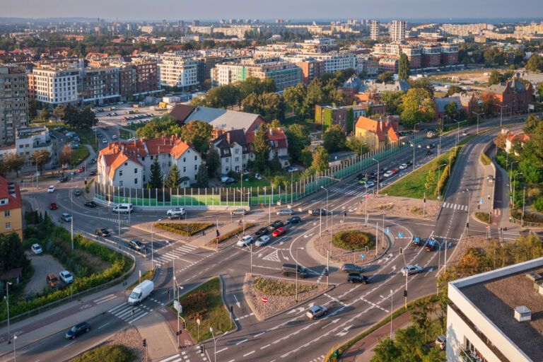 Widok z lotu ptaka na ruchliwe rondo otoczone budynkami, drzewami i zaparkowanymi samochodami w miejskiej dzielnicy Gdańska przy bezchmurnym niebie. Wokół ronda biegnie kilka pasów ruchu, po których poruszają się pojazdy wjeżdżające i wyjeżdżające.