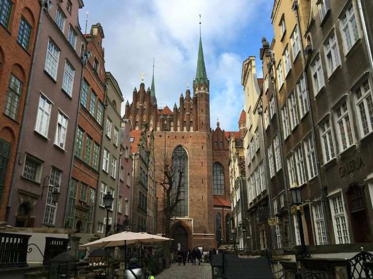 A cobblestone street lined with tall, historic buildings leads to a large Gothic church with a tall green spire, arched windows, and brick façade under a partly cloudy sky. Outdoor seating is visible along the pavement.