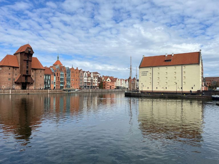 A calm river reflects historic brick and white buildings with red roofs under a partly cloudy sky in a Gdansk, Poland, with boats moored along the waterfront.