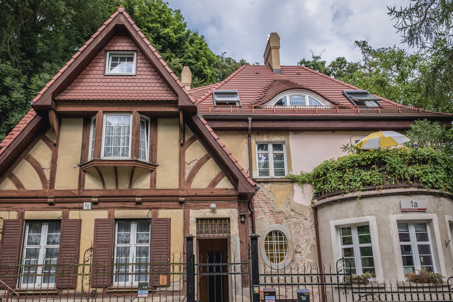 Uroczy dwupiętrowy dom z czerwoną dachówką, elementami szachulcowymi i wysokimi oknami. Po jednej stronie pnie się bluszcz, a na niewielkim balkonie widać parasol w żółte paski. W tle widać drzewa i chmury.