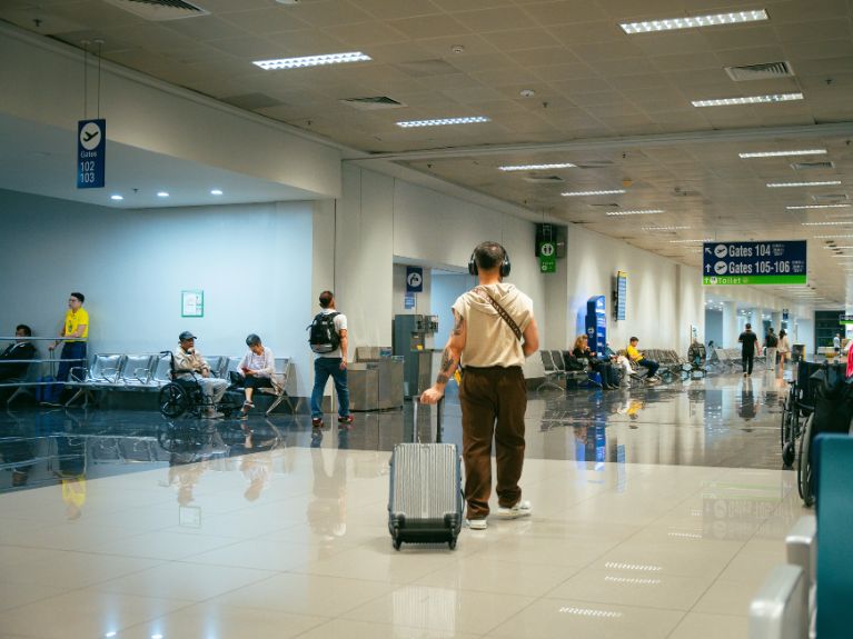 A person walks through an airport terminal pulling a suitcase. Other travellers sit on benches or walk, and gate signs are visible overhead. The area is brightly lit with a shiny tiled floor.