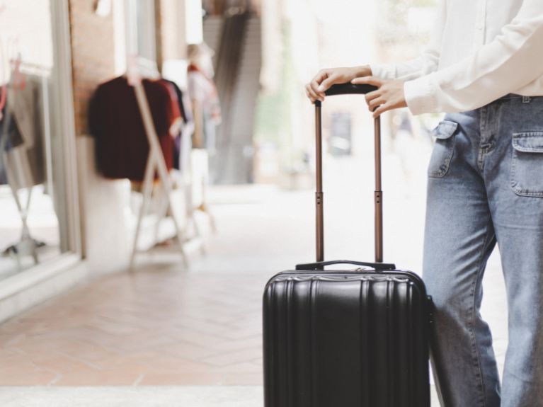 A person wearing blue jeans and a white shirt stands outdoors, holding the handle of a black rolling suitcase. Shopfronts and clothing displays are visible in the blurred background.