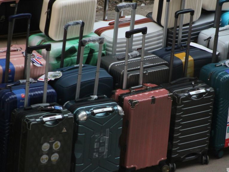 Several hard-shell suitcases in various colours and sizes are standing upright with extended handles, arranged closely together in rows, suggesting a busy travel or luggage storage area.