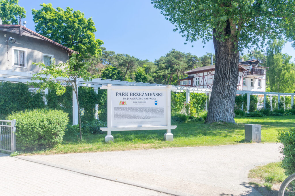 Duży napis „Park Brzeźnieński im. Jana Jerzego Haffnera" stoi na trawiastym terenie przy chodniku, w tle drzewa, zieleń i budynki mieszkalne w słoneczny dzień.