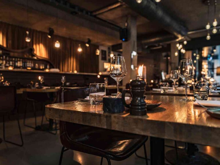 A dimly lit restaurant with wooden tables set with glasses, plates, and a lit candle. The background shows a bar area, warm lighting, and empty chairs, creating a cosy and inviting atmosphere.