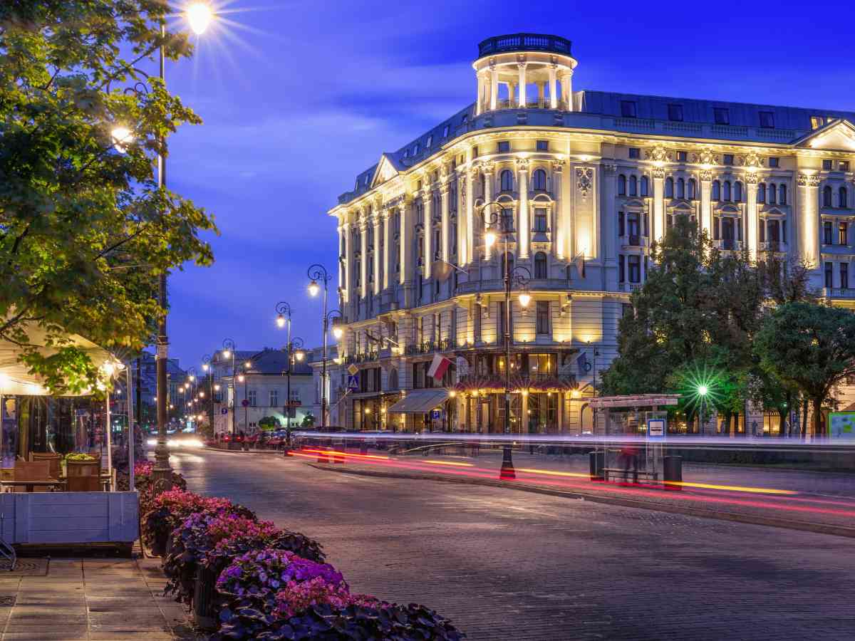 A grand, historic building with ornate lights glows at dusk on a city street. Trees and flowers line the pavement, while car lights create streaks along the empty road under a deep blue sky.