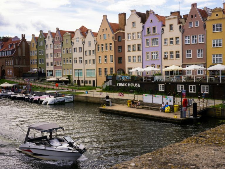 A row of colourful, historic buildings with outdoor seating lines a waterfront. A small boat moves along the river, and docks with people and moored boats are visible in the foreground.