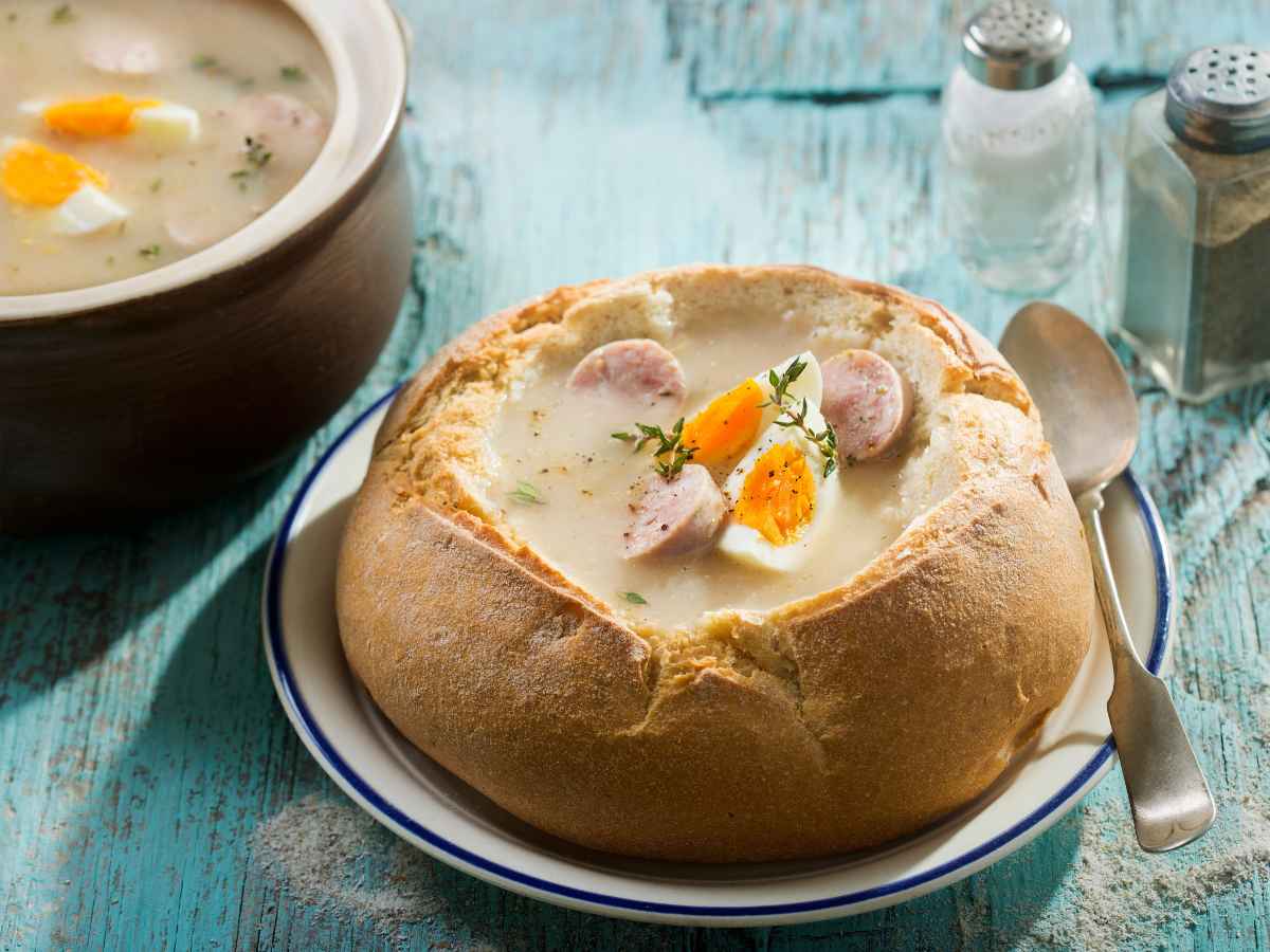 A bread bowl filled with creamy soup, garnished with sliced sausage, hard-boiled egg pieces, and fresh herbs, sits on a plate with a spoon. A pot of soup and salt and pepper shakers are in the background on a blue wooden table.