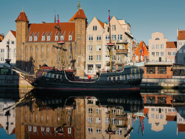 A historic-style tall ship with black sails and flags is moored on a calm river, reflecting old brick and pastel buildings under a clear blue sky.