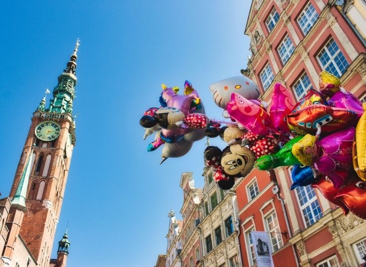 Kilka kolorowych balonów, w tym Myszka Miki, Hello Kitty i Świnka Peppa, unosi się przed wysoką wieżą zegarową i czerwonymi budynkami pod czystym, błękitnym niebem w Gdańsku.