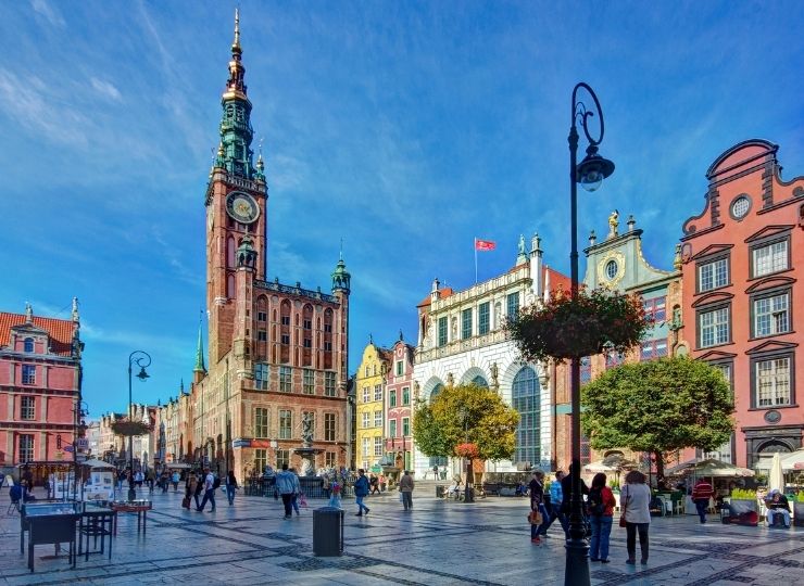 Ludzie spacerują szeroką, brukowaną ulicą wyłożoną kolorowymi zabytkowymi budynkami i wysoką wieżą zegarową pod jasnym, błękitnym niebem. Ozdobne lampy i drzewa dodają uroku tej tętniącej życiem miejskiej scenerii.