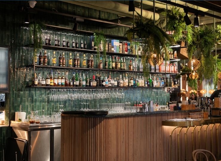 A stylish bar with green tiled walls, shelves stocked with bottles of alcohol and glasses, hanging plants, and warm pendant lighting creating a modern, inviting atmosphere.