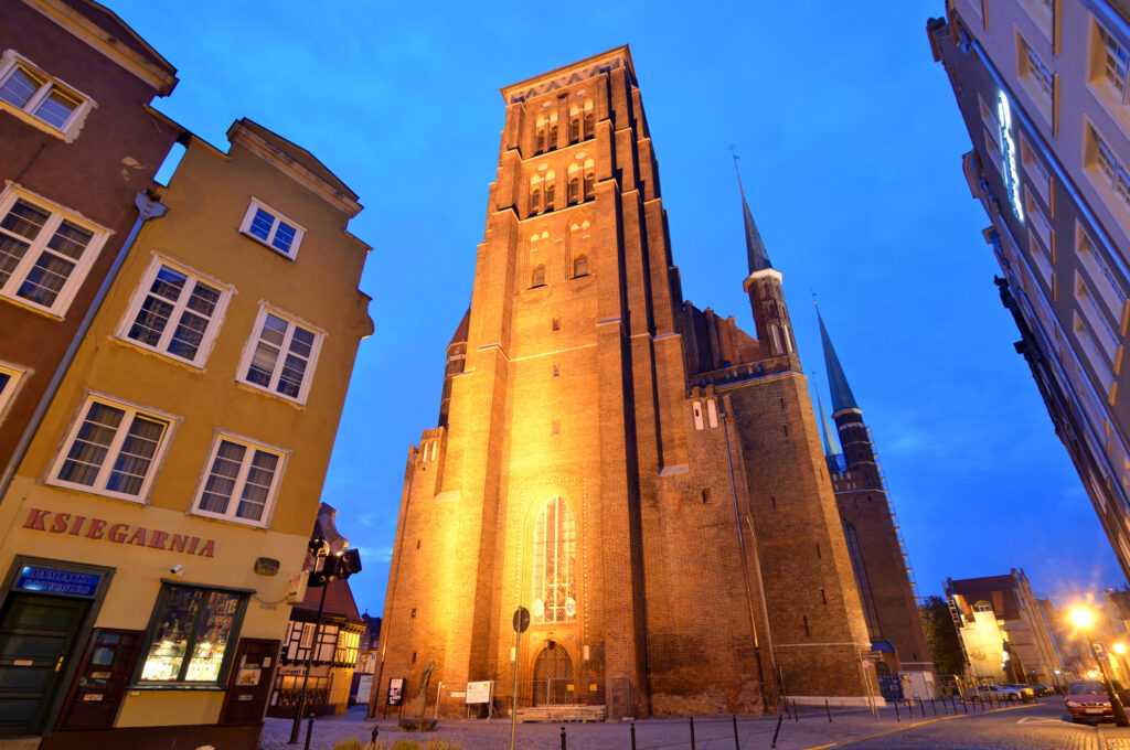 A tall, illuminated brick church with twin spires stands in a city square at dusk, surrounded by old buildings, including one with a sign reading KSIEGARNIA. The sky is a deep blue.