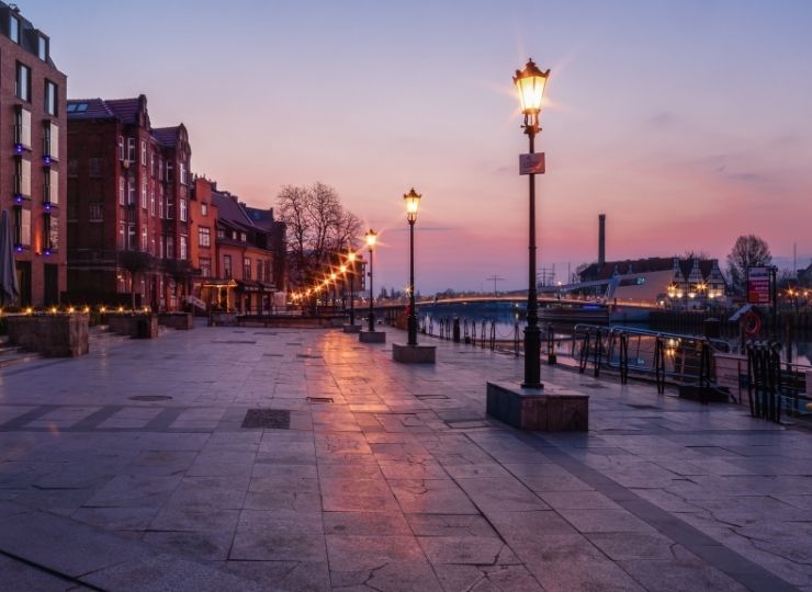 Cicha, pusta nadrzeczna promenada o zachodzie słońca, wzdłuż której stoją latarnie w starym stylu i historyczne budynki po lewej stronie, w tle spokojne wody i delikatnie różowo-fioletowe niebo.