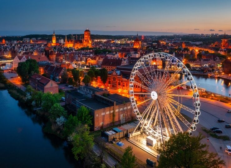 A large, illuminated Ferris wheel stands near a river at dusk, with a cityscape of historic buildings and softly glowing streetlights in the background. The sky is blue and orange as night falls.