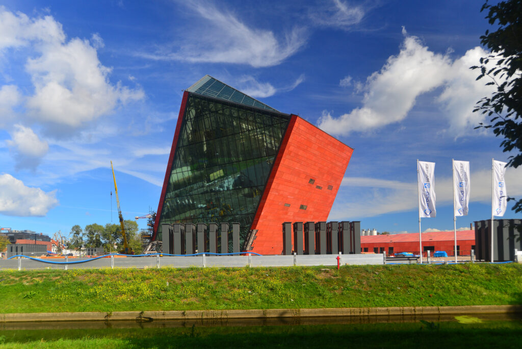 A modern, red, angular building with large glass windows stands against a blue sky with clouds, surrounded by grass and flags.