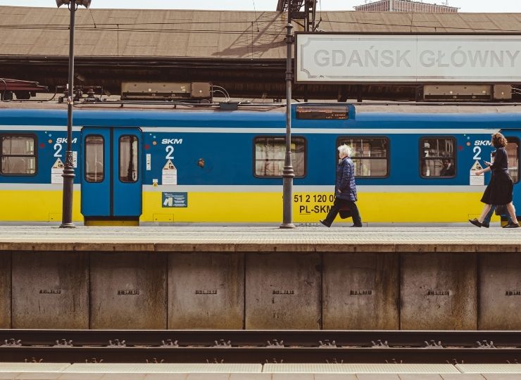 A blue and yellow train is stopped at the Gdańsk Główny station. Two women are walking on the platform in opposite directions, and the station sign is partially visible above the train.