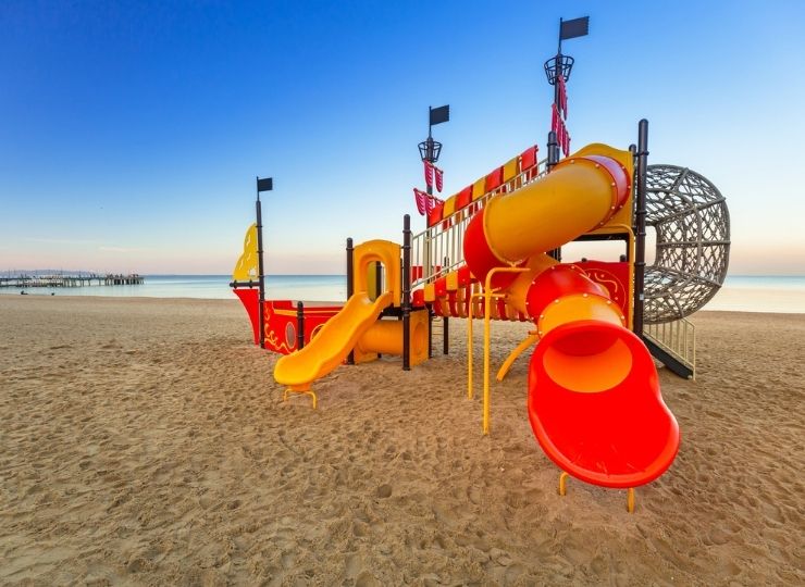Kolorowy plac zabaw w kształcie pirackiego statku ze zjeżdżalniami i siatkami do wspinaczki stoi na piaszczystej plaży, w tle spokojne morze i bezchmurne niebo.