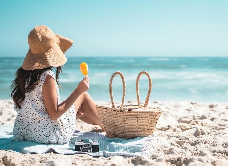 A woman in a sunhat and summer dress sits on a beach blanket, eating an ice lolly. A wicker basket and a camera are beside her on the sand, with the sea and blue sky in the background.
