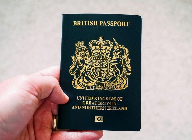 A hand holds a British passport with a dark cover featuring the UK coat of arms and the words “United Kingdom of Great Britain and Northern Ireland” in gold lettering.