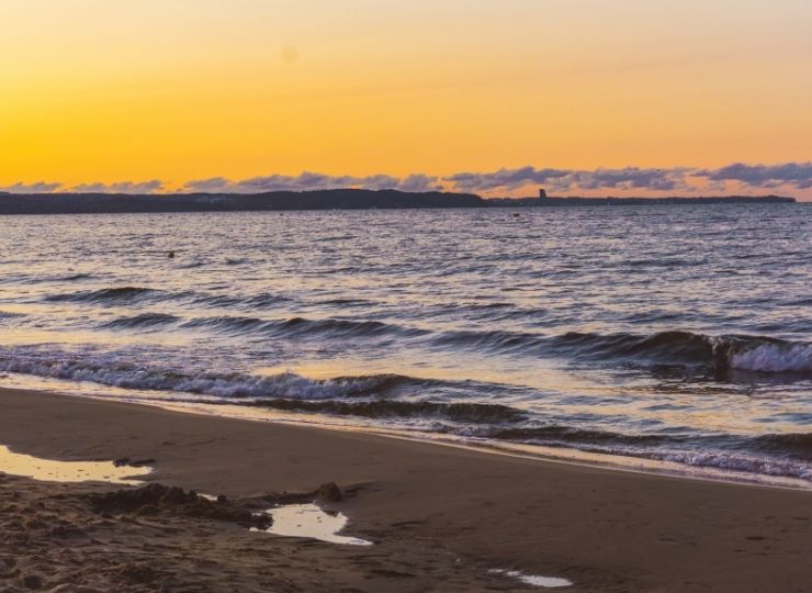 A peaceful beach scene at sunset, with gentle waves rolling onto the sandy shore and a distant coastline visible under an orange and yellow sky.