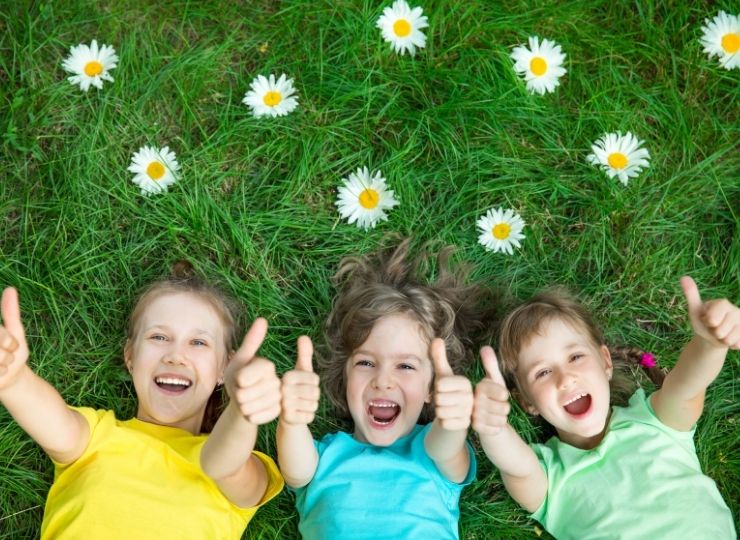 Three smiling children lying on green grass surrounded by daisies, all giving a thumbs-up gesture and wearing brightly coloured shirts.