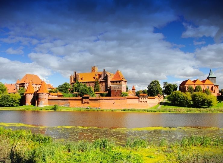 A large medieval red-brick castle with multiple towers and walls sits beside a river under a dramatic sky with dark and white clouds, surrounded by green trees and grass.