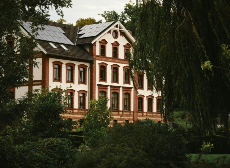 A large, three-storey brick and white house with solar panels on the roof, surrounded by lush greenery and trees.