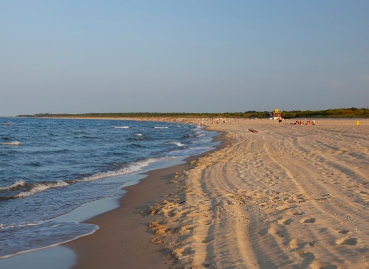 A sandy beach with gentle sea waves on the left and footprints in the sand. A few people and a lifeguard hut are visible in the distance under a clear blue sky.