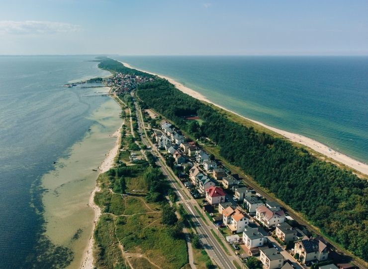 Aerial view of a narrow strip of land with houses, a road, dense greenery, and sandy beaches on both sides, bordered by blue sea under a clear sky.