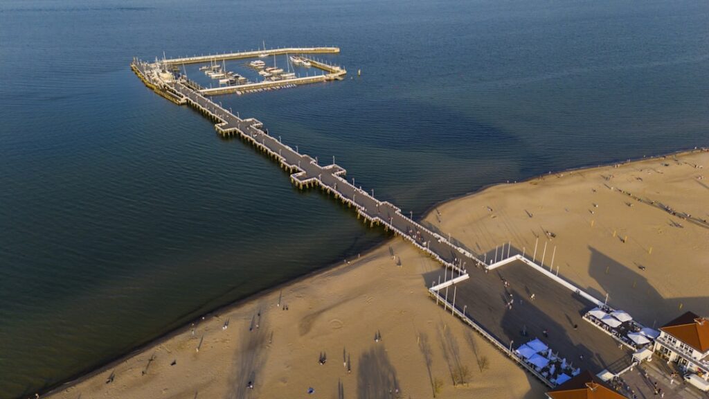 Aerial view of a long pier extending from a sandy beach into calm blue water, with boats moored at the end and a few people walking along the pier and shoreline.