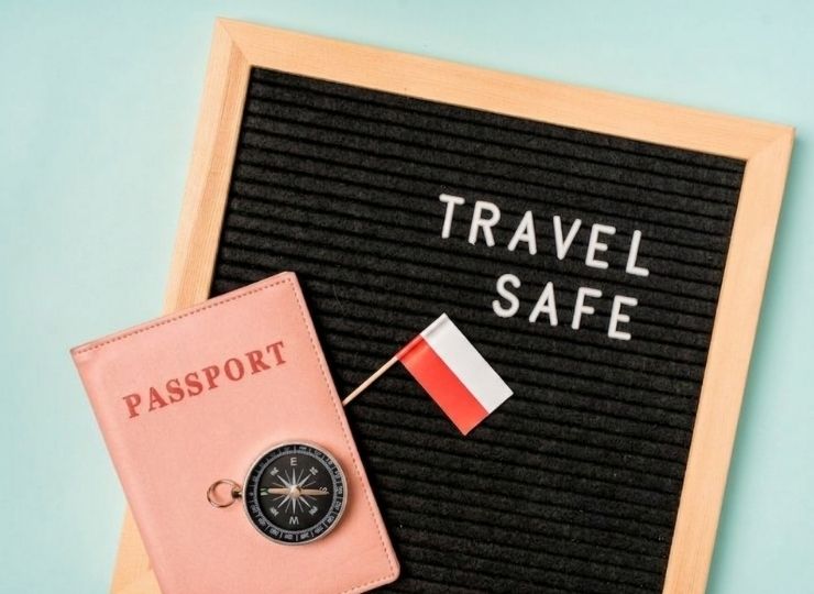 A pink passport, small compass, and Polish flag rest on a black felt letter board with the words TRAVEL SAFE on a light blue background.