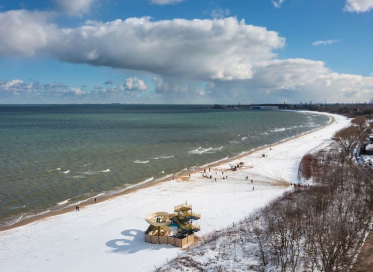 Aerial view of a snowy beach with a winding yellow waterslide, gentle waves, scattered people, and a partly cloudy sky. Trees line the right side and industrial structures are visible in the distant background.