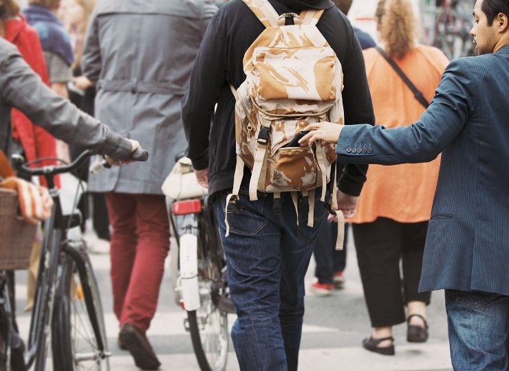 A person in a crowd is being pickpocketed; someone in a blue jacket is reaching into the rucksack of a person wearing dark jeans and a tan rucksack while others walk and ride bicycles nearby.