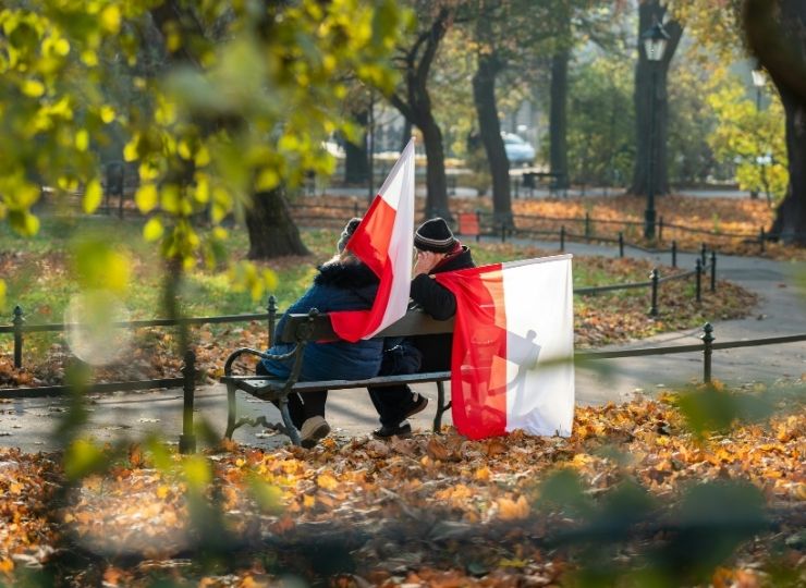 Two people sit on a park bench in autumn, holding and draping Polish flags. Fallen leaves cover the ground and trees with autumn foliage surround them, creating a peaceful scene.