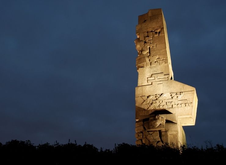 A tall, stone monument with abstract carvings is illuminated against a dark, cloudy evening sky, with the base surrounded by low vegetation.
