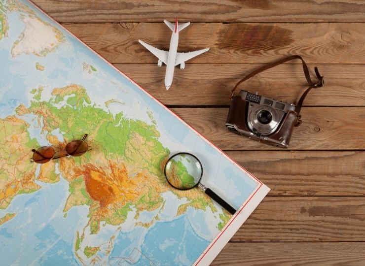 A map on a wooden table with a pair of sunglasses, a magnifying glass, a vintage camera with a strap, and a model aeroplane placed around it, suggesting travel planning.