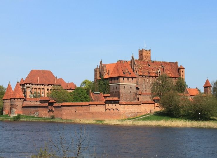 A large medieval brick castle with red roofs sits beside a river under a clear blue sky, surrounded by trees and greenery.