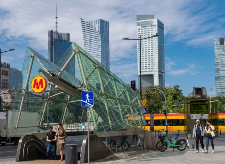 People walk near a modern glass metro entrance with skyscrapers in the background, bicycles parked nearby, and a tram passing by on a sunny day in a city.