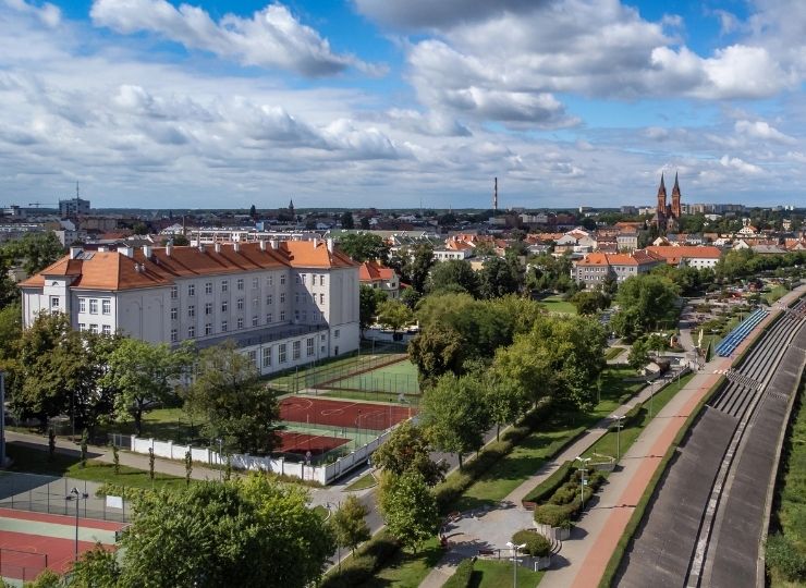 Aerial view of a cityscape with a large building, tennis courts, green trees, walking paths, and distant buildings under a partly cloudy sky.