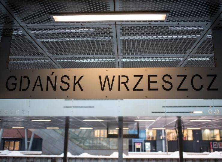 Табличка з написом GDAŃSK WRZESZCZ висить на сучасному критому залізничному вокзалі зі скляними стінами та засніженою землею зовні.